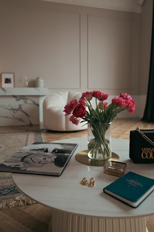 A chic, elegantly furnished living room with a round marble coffee table adorned with various items such as a fashion magazine, a small notebook, a glass perfume bottle, and a couple of rings. A vase filled with vibrant pink tulips adds a pop of color to the subdued color scheme. In the background, there is a plush white armchair and a white marble console table with decorative items.