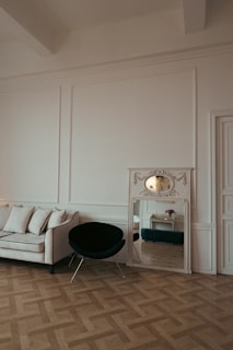A minimalist interior features a white sofa with black piping, a black chair with metal legs, and a decorative mirror placed on the floor. The room has ornate molding and a parquet wooden floor. There is a reflection in the mirror showing a small table with a flower vase.