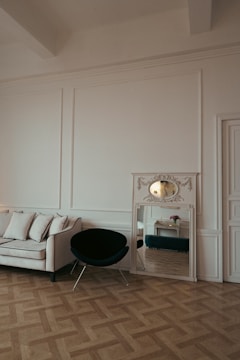 A minimalist interior features a white sofa with black piping, a black chair with metal legs, and a decorative mirror placed on the floor. The room has ornate molding and a parquet wooden floor. There is a reflection in the mirror showing a small table with a flower vase.