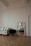 A minimalist interior features a white sofa with black piping, a black chair with metal legs, and a decorative mirror placed on the floor. The room has ornate molding and a parquet wooden floor. There is a reflection in the mirror showing a small table with a flower vase.