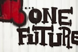 Graffiti art featuring the phrase 'One Future' painted in bold, black letters on a white, corrugated metal surface. A large red symbol resembling a fist or heart is present on the left side of the text.