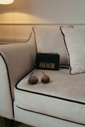 A luxurious setting is accentuated with a plush, cream-colored sofa featuring black piping on its edges. A stylish black designer bag with gold lettering and accents rests against the cushions, lending a touch of elegance. In the foreground, a pair of chic sunglasses with tinted lenses is casually placed on the sofa.