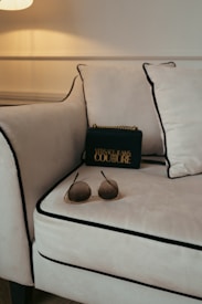 A luxurious setting is accentuated with a plush, cream-colored sofa featuring black piping on its edges. A stylish black designer bag with gold lettering and accents rests against the cushions, lending a touch of elegance. In the foreground, a pair of chic sunglasses with tinted lenses is casually placed on the sofa.