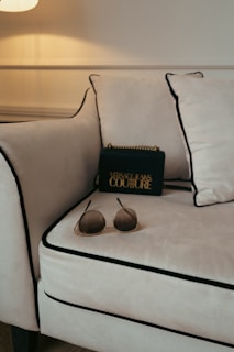 A luxurious setting is accentuated with a plush, cream-colored sofa featuring black piping on its edges. A stylish black designer bag with gold lettering and accents rests against the cushions, lending a touch of elegance. In the foreground, a pair of chic sunglasses with tinted lenses is casually placed on the sofa.