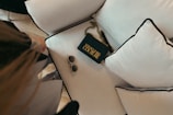 An elegant setting featuring a black handbag with gold chain detailing and bold lettering next to a pair of stylish sunglasses. The items are placed on a plush white cushion with black piping that rests on a light-colored wooden floor.