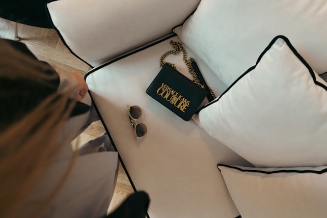 An elegant setting featuring a black handbag with gold chain detailing and bold lettering next to a pair of stylish sunglasses. The items are placed on a plush white cushion with black piping that rests on a light-colored wooden floor.