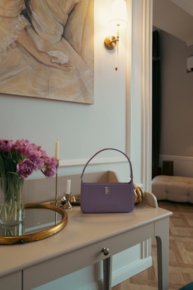 A well-arranged table featuring a lilac handbag placed next to a vase of pink flowers and a couple of candles. Above the table is a wall-mounted artwork depicting an embrace, and a wall sconce with a light provides soft illumination. The background includes a plush ottoman and a portion of an elegantly designed room with neutral tones.
