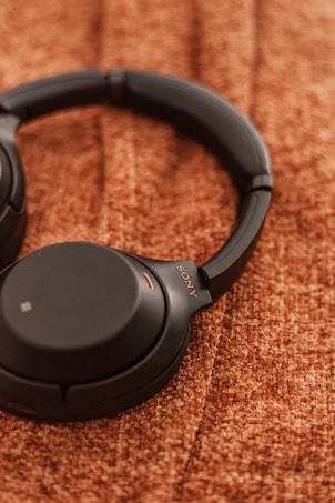 A pair of black over-ear headphones lies on a textured brown surface. The headphones have a sleek design with cushioned ear cups and a visible brand logo printed in gold.