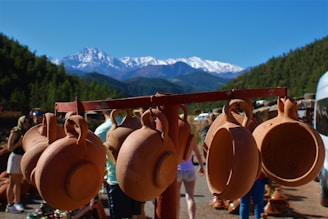 Clay pots are hanging on a wooden rod against a backdrop of scenic mountains. A group of people is gathered nearby, with a clear blue sky above and lush green trees surrounding the area. Snow-capped peaks are visible in the distance.