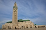A grand mosque with a tall minaret featuring intricate geometric designs dominates the scene. The architecture is a blend of traditional Islamic elements and modern touches with a predominantly beige facade and green tiled roofs. A wide open courtyard surrounds the mosque with several people walking around.