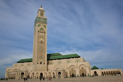 A grand mosque with a tall minaret featuring intricate geometric designs dominates the scene. The architecture is a blend of traditional Islamic elements and modern touches with a predominantly beige facade and green tiled roofs. A wide open courtyard surrounds the mosque with several people walking around.