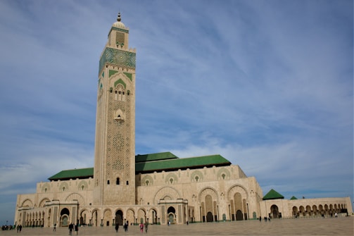 A grand mosque with a tall minaret featuring intricate geometric designs dominates the scene. The architecture is a blend of traditional Islamic elements and modern touches with a predominantly beige facade and green tiled roofs. A wide open courtyard surrounds the mosque with several people walking around.