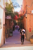 A narrow alleyway is lined with vibrant orange buildings and lush palm trees. Several people walk down the cobblestone path, and a sign on the left reads 'Les Bains d'Orient spa'. The alley is adorned with tangled electrical wires, and there is a bicycle parked against the wall.