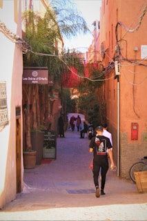 A narrow alleyway is lined with vibrant orange buildings and lush palm trees. Several people walk down the cobblestone path, and a sign on the left reads 'Les Bains d'Orient spa'. The alley is adorned with tangled electrical wires, and there is a bicycle parked against the wall.