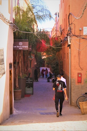 A narrow alleyway is lined with vibrant orange buildings and lush palm trees. Several people walk down the cobblestone path, and a sign on the left reads 'Les Bains d'Orient spa'. The alley is adorned with tangled electrical wires, and there is a bicycle parked against the wall.