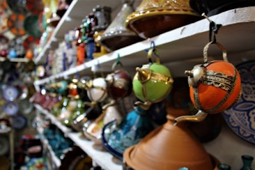 A display of colorful, intricately designed pottery and decorative items on shelves, featuring various shapes and ornamental patterns. The focus is on ornate teapots and bowls with vibrant hues.