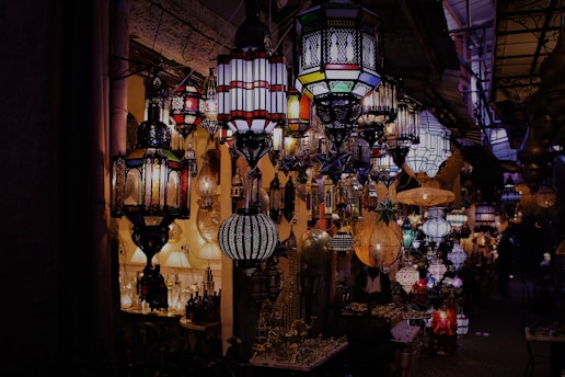 A vibrant and colorful array of intricately designed lamps and lanterns displayed in a dimly lit marketplace, creating an enchanting and exotic ambiance. The variety of shapes and patterns cast intricate shadows and reflections on the surrounding surfaces.