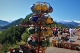 A display of colorful ceramic mugs is the focal point, set against a scenic backdrop of lush green hills and distant snow-capped mountains. In the background, people are browsing various pottery items laid out on tables, suggesting an outdoor market setting under a clear blue sky.