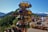 A display of colorful ceramic mugs is the focal point, set against a scenic backdrop of lush green hills and distant snow-capped mountains. In the background, people are browsing various pottery items laid out on tables, suggesting an outdoor market setting under a clear blue sky.