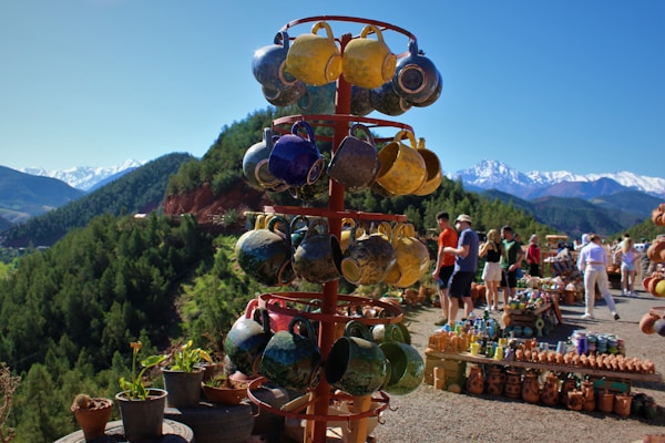 A display of colorful ceramic mugs is the focal point, set against a scenic backdrop of lush green hills and distant snow-capped mountains. In the background, people are browsing various pottery items laid out on tables, suggesting an outdoor market setting under a clear blue sky.