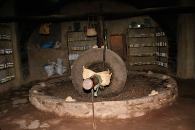 An interior setting featuring a large, stone grinding wheel in the center of a circular pit. Surrounding the wheel are rustic stone walls lined with shelves holding numerous bottles. Aesthetic elements like bags and baskets add to the atmosphere, suggesting possible storage or production purposes. The lighting is dim, emphasizing the rough textures of the stone and walls.