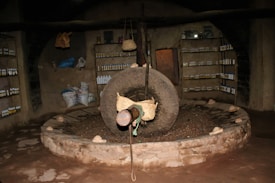 An interior setting featuring a large, stone grinding wheel in the center of a circular pit. Surrounding the wheel are rustic stone walls lined with shelves holding numerous bottles. Aesthetic elements like bags and baskets add to the atmosphere, suggesting possible storage or production purposes. The lighting is dim, emphasizing the rough textures of the stone and walls.