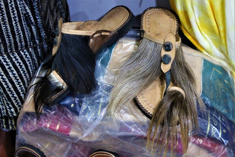 A pair of traditional leather sandals with long hair attached is displayed. They rest on a clear plastic cover sheet, and various fabrics and items can be seen underneath the plastic. The sandals have intricate details and stitching, with the hair varying in color from black to light brown. There is a yellow and white fabric on the right side and a black and white patterned fabric on the left side.