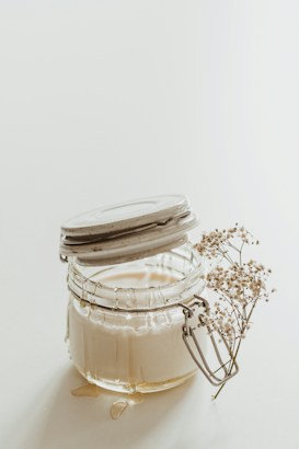 A glass jar filled with a creamy substance is placed against a light background. The jar is open, with its lid tilted, and some of the cream has spilled onto the surface. Attached to the jar is a sprig of delicate baby's breath flowers.
