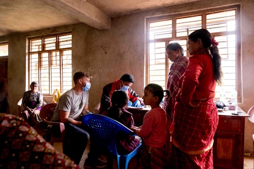 A group of people is gathered inside a dimly lit room with large windows. A medical professional wearing a mask is seated and attending to a child while others, including women and an elderly person, are either seated or standing nearby. The interior has plain walls, and there appears to be a consultation taking place.