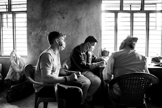 Three individuals are seated in a dimly lit room with barred windows. Two of them are facing away from the camera while one is visible in profile. They all appear to be engaged in a discussion or examination, and are wearing face masks. The room has a rough, unfinished look with some bags and other items placed on the floor and a chalkboard scribbled with writing on the wall.