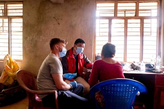Three people are seated indoors near a window, all wearing masks. The room has a simple, unfinished appearance with bare walls and minimal furniture. One person appears to be taking notes or filling out a form as they interact with the others. A plastic chair and various items rest on a cluttered table.