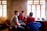Three people are seated indoors near a window, all wearing masks. The room has a simple, unfinished appearance with bare walls and minimal furniture. One person appears to be taking notes or filling out a form as they interact with the others. A plastic chair and various items rest on a cluttered table.