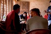 Several people are gathered in a dimly lit room. Most of them are wearing masks and having a serious conversation. Some individuals in the background are seated on chairs, while others are engaged in discussion around a table. The room has a rustic appearance with wooden windows and doors.