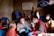 Several people are gathered in a room with basic furnishings. A man in a grey shirt and blue gloves is engaging with an elderly woman wearing a red patterned garment, possibly discussing paperwork. Others in the background are seated or standing, some wearing face masks. The room contains blue plastic chairs, and there are posters or signs with writing on the walls.