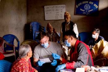 Several people are gathered in a room with basic furnishings. A man in a grey shirt and blue gloves is engaging with an elderly woman wearing a red patterned garment, possibly discussing paperwork. Others in the background are seated or standing, some wearing face masks. The room contains blue plastic chairs, and there are posters or signs with writing on the walls.