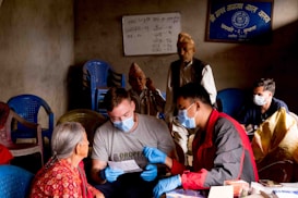 Several people are gathered in a room with basic furnishings. A man in a grey shirt and blue gloves is engaging with an elderly woman wearing a red patterned garment, possibly discussing paperwork. Others in the background are seated or standing, some wearing face masks. The room contains blue plastic chairs, and there are posters or signs with writing on the walls.