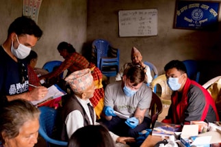 A group of people, some wearing masks, are gathered in a room with blue plastic chairs. One person is taking notes while others appear to be engaged in a conversation or medical examination. A board with writing is visible on the wall.