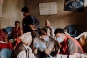 A group of people are gathered in a room, with some seated on blue plastic chairs. Two men in the foreground are wearing face masks and engaging in conversation, one holding a syringe, hinting at a medical setting. An older man wearing a traditional Nepali hat and vest is seated, conversing with the men. A woman in a colorful traditional dress looks on attentively from her chair. The walls have written notices and a framed sign in a non-English script. The setting appears based on community health outreach or vaccination drive.
