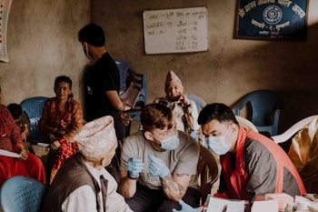 A group of people are gathered in a room, with some seated on blue plastic chairs. Two men in the foreground are wearing face masks and engaging in conversation, one holding a syringe, hinting at a medical setting. An older man wearing a traditional Nepali hat and vest is seated, conversing with the men. A woman in a colorful traditional dress looks on attentively from her chair. The walls have written notices and a framed sign in a non-English script. The setting appears based on community health outreach or vaccination drive.
