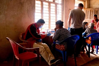 A group of people are gathered inside a room with muted light filtering through a window. A person wearing a face mask and a red jacket sits on a plastic chair while interacting with a seated woman. Other individuals in the room are engaged in conversations and appear to be involved in a community activity. The walls are bare and some writing is visible. The setting gives the impression of an informal communal space.