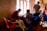 A group of people are gathered inside a room with muted light filtering through a window. A person wearing a face mask and a red jacket sits on a plastic chair while interacting with a seated woman. Other individuals in the room are engaged in conversations and appear to be involved in a community activity. The walls are bare and some writing is visible. The setting gives the impression of an informal communal space.