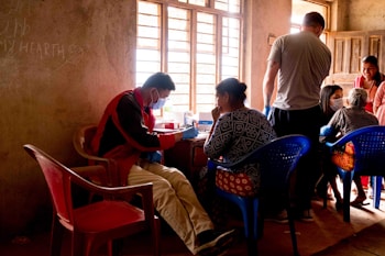 A group of people are gathered inside a room with muted light filtering through a window. A person wearing a face mask and a red jacket sits on a plastic chair while interacting with a seated woman. Other individuals in the room are engaged in conversations and appear to be involved in a community activity. The walls are bare and some writing is visible. The setting gives the impression of an informal communal space.
