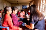 A room filled with several people, with a focus on a woman and child seated on blue chairs. The woman, wearing a red outfit, watches as the child receives a check-up from a person with long hair, possibly a healthcare worker, who holds medical equipment. In the background, more individuals, some with medical masks, are engaged in activities, likely related to health services. Posters and a blue banner adorn the walls, and sunlight streams through barred windows.