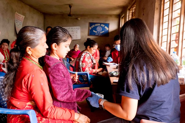 A room filled with several people, with a focus on a woman and child seated on blue chairs. The woman, wearing a red outfit, watches as the child receives a check-up from a person with long hair, possibly a healthcare worker, who holds medical equipment. In the background, more individuals, some with medical masks, are engaged in activities, likely related to health services. Posters and a blue banner adorn the walls, and sunlight streams through barred windows.