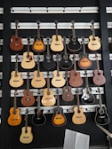 A variety of acoustic guitars are displayed on a black wall with horizontal white slats. The guitars have different shapes, sizes, and colors, including black, brown, natural wood, and sunburst finishes. At the bottom right corner, there is a partially visible open book with musical notes.