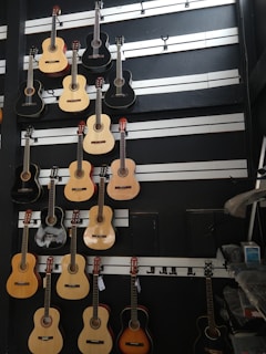 A wall displaying various acoustic guitars arranged in rows. The guitars differ in color and design, including shades of natural wood, black, and sunburst. Each guitar hangs on hooks attached to horizontal strips. The background is dark with white strips horizontally aligned, enhancing the visibility of the guitars.