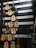 A wall displaying various acoustic guitars arranged in rows. The guitars differ in color and design, including shades of natural wood, black, and sunburst. Each guitar hangs on hooks attached to horizontal strips. The background is dark with white strips horizontally aligned, enhancing the visibility of the guitars.