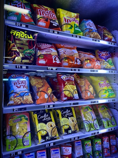 A vending machine filled with a variety of snacks and beverages, including bags of chips and packs of cookies and drinks. The packaging features bright colors and a mix of English and another script, likely Thai.