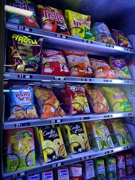 A vending machine filled with a variety of snacks and beverages, including bags of chips and packs of cookies and drinks. The packaging features bright colors and a mix of English and another script, likely Thai.