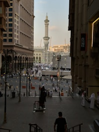 A bustling cityscape featuring a large, ornate mosque tower with detailed architecture, surrounded by high-rise buildings. The street is filled with people, many in traditional attire, suggesting a busy urban environment.